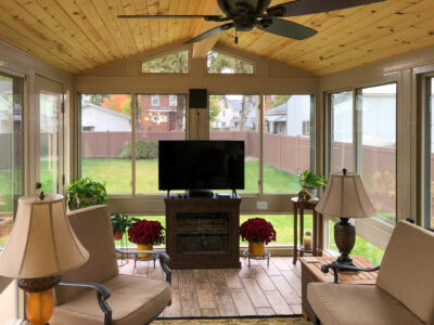 Decorated sunroom with natural wood ceiling and rustic wood plank tile floor. The room features two cozy beige armchairs, a central fireplace media console with a large TV, and tall windows offering a view of the green backyard. The space is accessorized with large table lamps, potted plants, and red chrysanthemum decor, showing a comfortable all-season room design ready for relaxation.