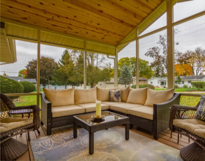 Interior view of a custom screened porch addition in Southern Wisconsin featuring a wicker sectional, a beautiful wood-plank ceiling, and a clear view of a grassy backyard. All while being protected from the pests and elements. This is just one of the benefits of a screen room in Wisconsin.