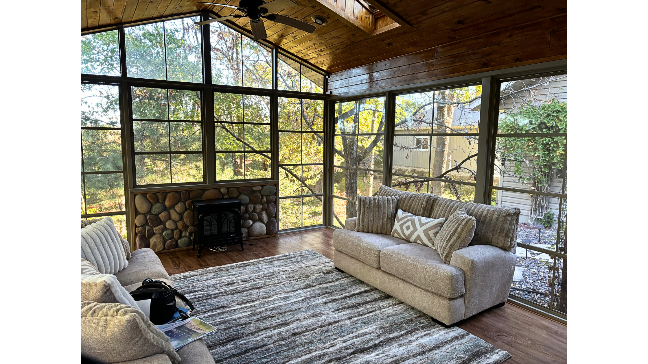 An affordable porch enclosure system featuring a cozy interior with comfortable sofas, a large area rug, a wood-paneled ceiling with a fan, and large windows overlooking trees.