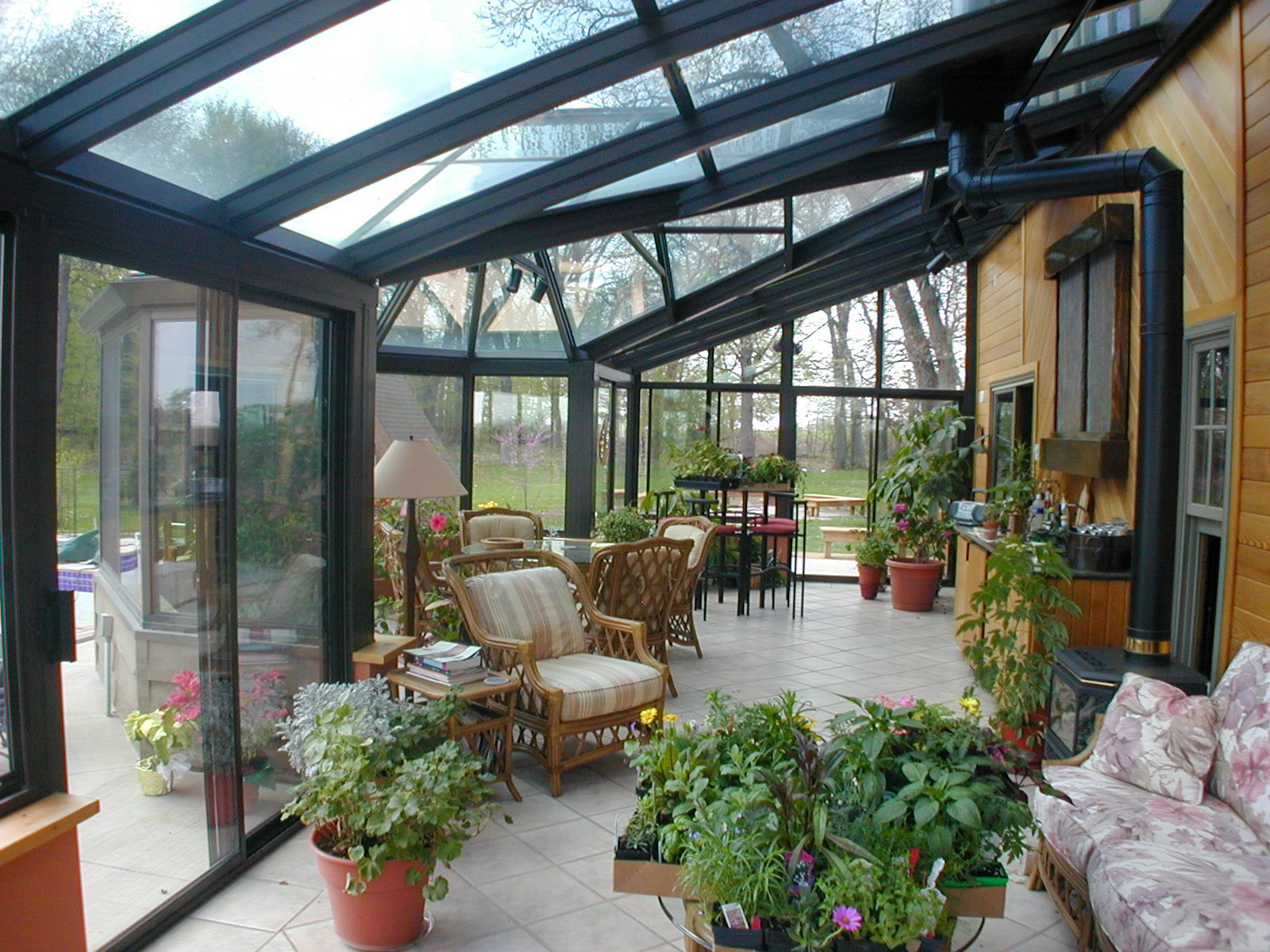 A glass-roof sunroom greenhouse setup filled with potted indoor plants, patio furniture, and tiled flooring overlooking a yard.