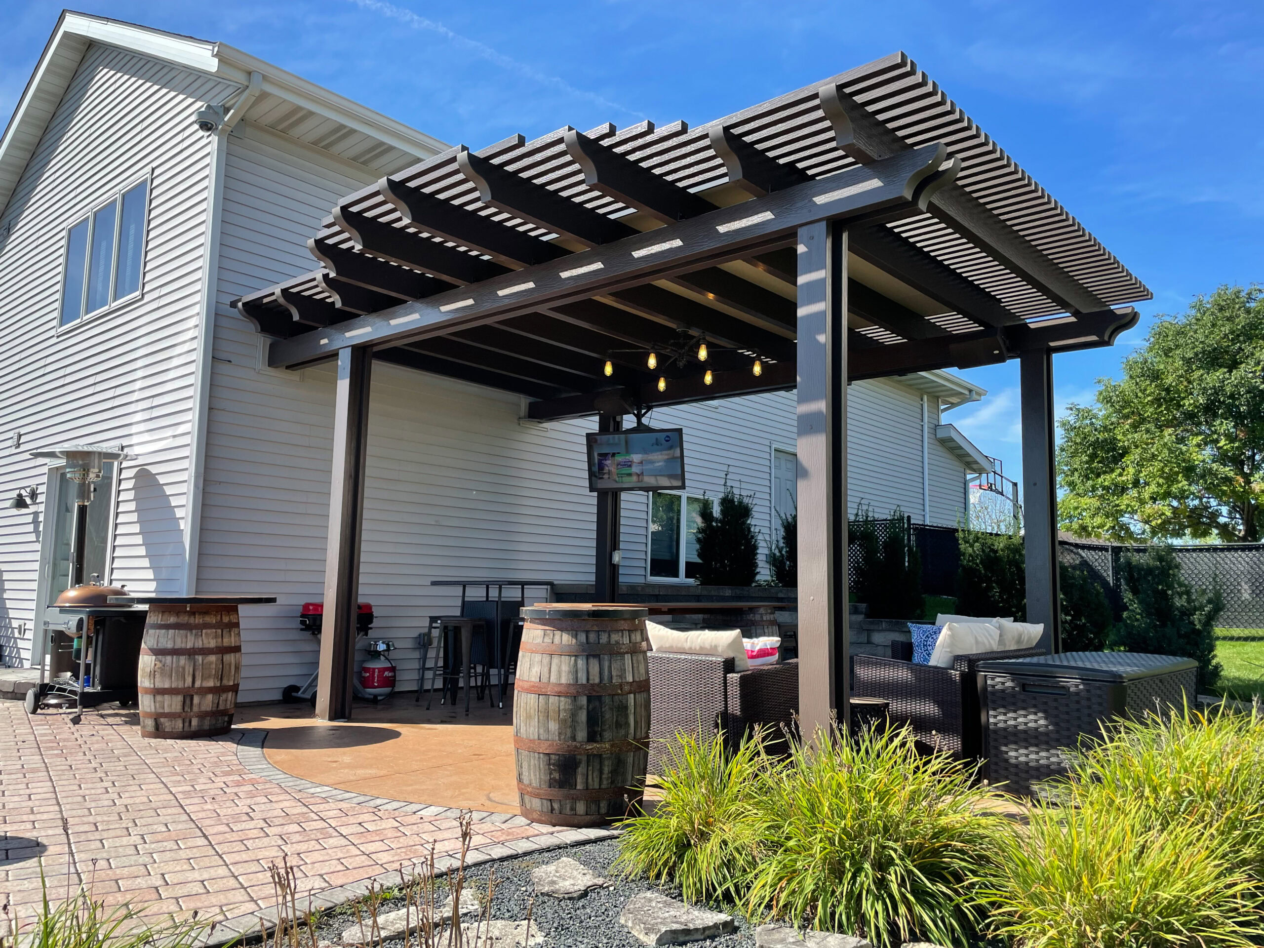Custom low-maintenance dark brown pergola installed over a stone patio with outdoor seating and television in Southern Wisconsin.