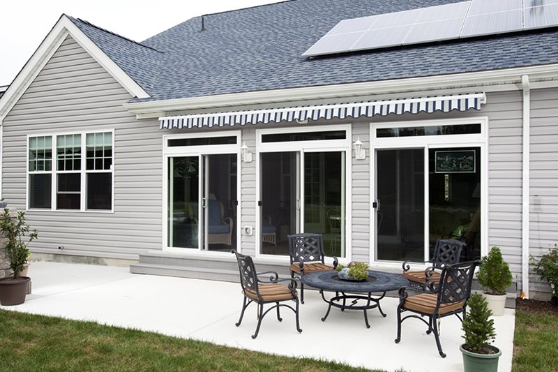 A detailed view of a partially retracted black and white striped motorized awning installed above two sliding glass patio doors on a modern gray-sided home, showing the cassette housing and compact design when not in use.