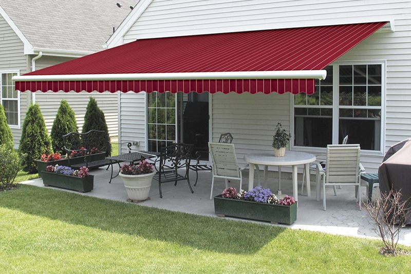 A spacious patio with seating and dining areas is shaded by a large, extended red motorized retractable awning. The awning fabric is a deep crimson color and provides cool coverage for the entire patio space.